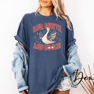 May include: Navy blue t-shirt with a distressed graphic that reads "Red, White, and Goose!" featuring a goose with wings designed as the American flag. The shirt is a relaxed fit.