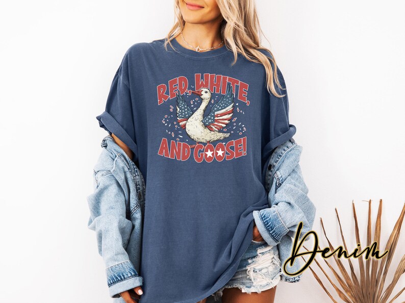 May include: Navy blue t-shirt with a distressed graphic that reads "Red, White, and Goose!" featuring a goose with wings designed as the American flag. The shirt is a relaxed fit.