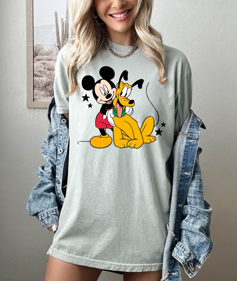 May include: A light green t-shirt with a cartoon image of Mickey Mouse and Pluto. Mickey Mouse is wearing red shorts and Pluto is wearing a green collar.