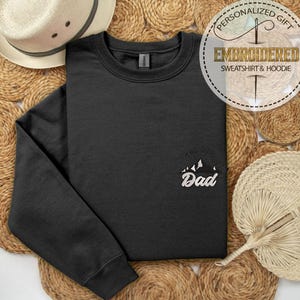 Op de afbeelding: Een zwart sweatshirt met het woord "Dad" in het wit geborduurd, samen met een bergafbeelding. Het sweatshirt wordt gepresenteerd op een geweven achtergrond, met een strohoed en een waaier in de afbeelding.