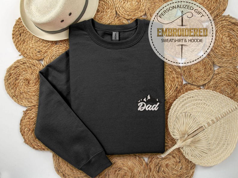 Op de afbeelding: Een zwart sweatshirt met het woord "Dad" in het wit geborduurd, samen met een bergafbeelding. Het sweatshirt wordt gepresenteerd op een geweven achtergrond, met een strohoed en een waaier in de afbeelding.