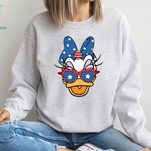 May include: A gray sweatshirt with a cartoon duck wearing red, white, and blue sunglasses and a bow with stars. The duck has a white face and a yellow beak.