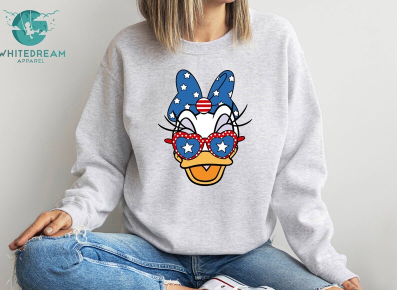 May include: A gray sweatshirt with a cartoon duck wearing red, white, and blue sunglasses and a bow with stars. The duck has a white face and a yellow beak.
