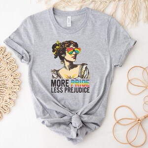 May include: Gray t-shirt with a colorful illustration of a woman wearing rainbow heart-shaped sunglasses. The text "More Pride Less Prejudice" is printed below the illustration.