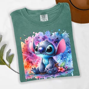 Pu&ograve; includere: T-shirt verde con un'illustrazione ad acquerello del personaggio Stitch di Lilo & Stitch. L'opera mostra Stitch con pelliccia blu, grandi occhi neri e orecchie rosa, su uno sfondo colorato a spruzzi. La maglietta &egrave; piegata.