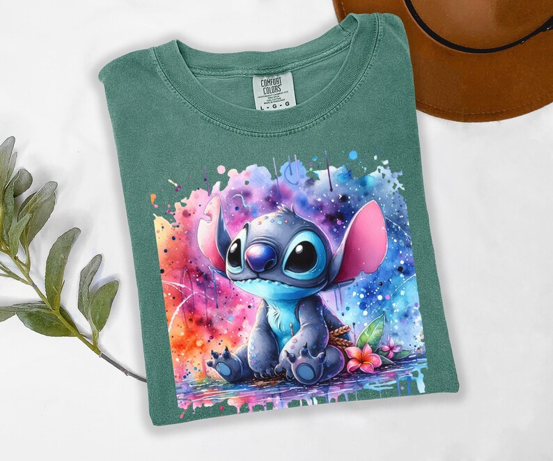 Pu&ograve; includere: T-shirt verde con un'illustrazione ad acquerello del personaggio Stitch di Lilo & Stitch. L'opera mostra Stitch con pelliccia blu, grandi occhi neri e orecchie rosa, su uno sfondo colorato a spruzzi. La maglietta &egrave; piegata.
