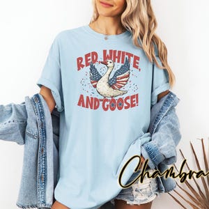 May include: Light blue t-shirt with a patriotic design. The graphic features a goose with wings styled as the American flag, accompanied by the text "Red, White, and Goose!" in red and white. The shirt is part of a casual clothing collection.