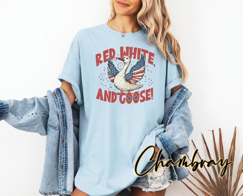 May include: Light blue t-shirt with a patriotic design. The graphic features a goose with wings styled as the American flag, accompanied by the text "Red, White, and Goose!" in red and white. The shirt is part of a casual clothing collection.