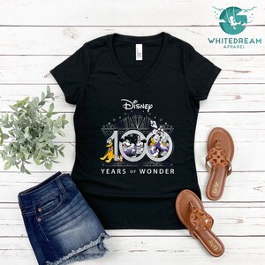 May include: Black V-neck t-shirt with a graphic of Mickey Mouse, Donald Duck, Goofy, and Pluto celebrating Disney's 100 Years of Wonder. The graphic is in silver and black with a diamond design.