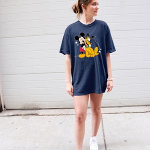 May include: A woman wearing a navy blue t-shirt with a cartoon image of Mickey Mouse and Pluto. The shirt has a relaxed fit and is worn with white high-top sneakers.