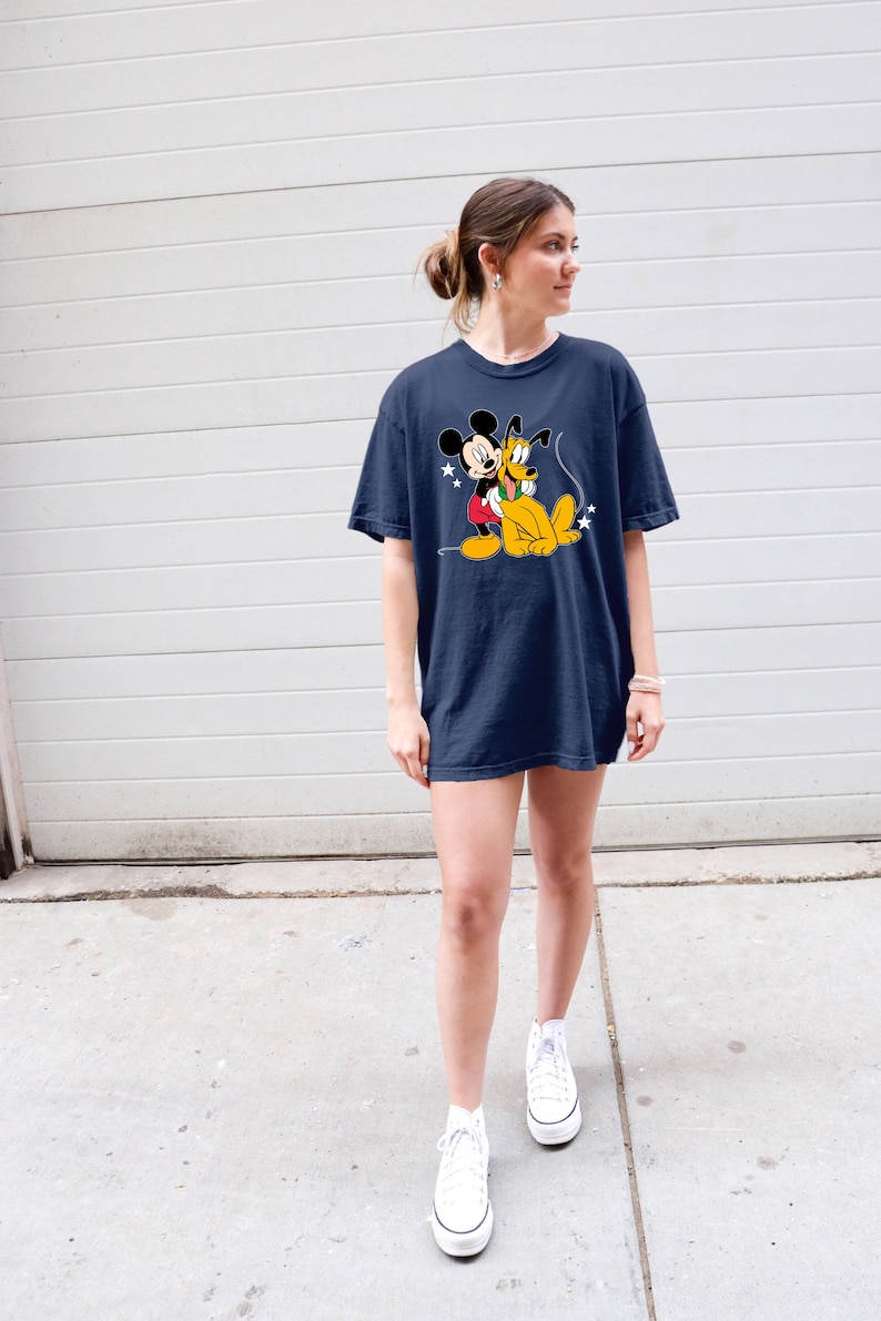 May include: A woman wearing a navy blue t-shirt with a cartoon image of Mickey Mouse and Pluto. The shirt has a relaxed fit and is worn with white high-top sneakers.