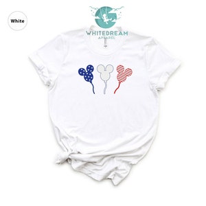 Puede incluir: Camiseta blanca con un gr&aacute;fico de tres globos con forma de Mickey Mouse. Los globos est&aacute;n decorados con rayas y estrellas rojas, blancas y azules, que recuerdan a la bandera espa&ntilde;ola.