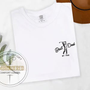 K&ouml;nnte beinhalten: Wei&szlig;es T-Shirt mit dem Text "Best Dad by Par" und einer Grafik eines Golfspielers. Das Shirt hat einen Rundhalsausschnitt und besteht aus Comfort Colors-Material. Ein runder Aufkleber zeigt "Personalized Gift" und "Embroidered Comfort Colors."