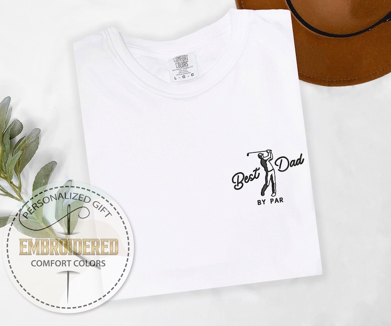 K&ouml;nnte beinhalten: Wei&szlig;es T-Shirt mit dem Text "Best Dad by Par" und einer Grafik eines Golfspielers. Das Shirt hat einen Rundhalsausschnitt und besteht aus Comfort Colors-Material. Ein runder Aufkleber zeigt "Personalized Gift" und "Embroidered Comfort Colors."
