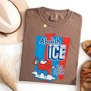 May include: Brown t-shirt with the words "Abolish ICE" and a cartoon polar bear holding a drink. The shirt also has the phrase "keep families together". A gold necklace, sandals, and a hat are also in the image.