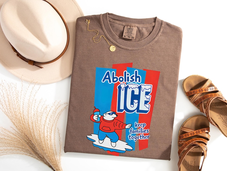 May include: Brown t-shirt with the words "Abolish ICE" and a cartoon polar bear holding a drink. The shirt also has the phrase "keep families together". A gold necklace, sandals, and a hat are also in the image.
