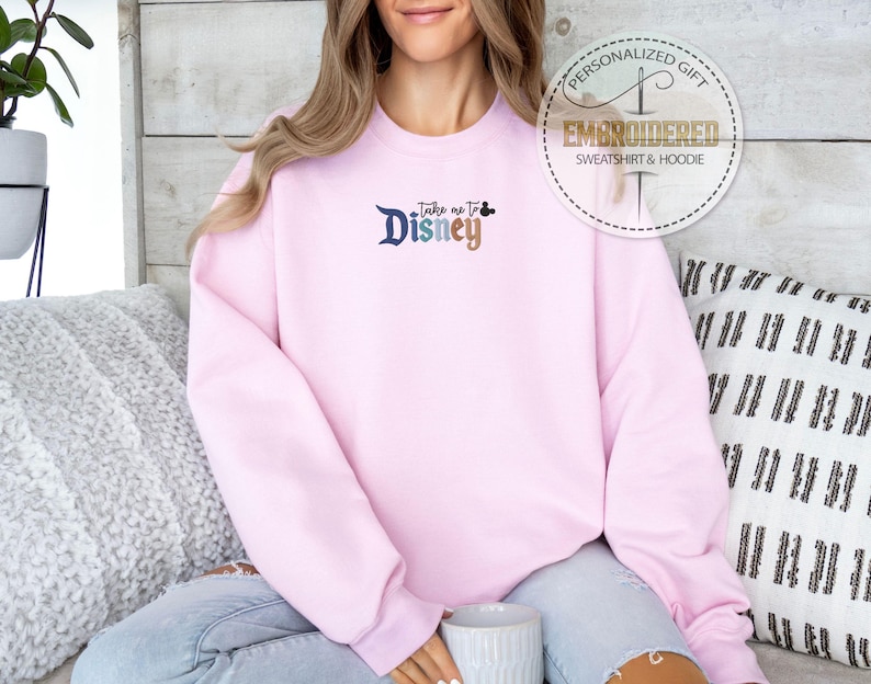 Puede incluir: Una sudadera rosa claro bordada con el texto "Take me to Disney" en azul y marr&oacute;n. La sudadera tiene cuello redondo y mangas largas. Un logotipo circular en la parte superior derecha dice "Personalized Gift" y "Embroidered Sweatshirt & Hoodie."