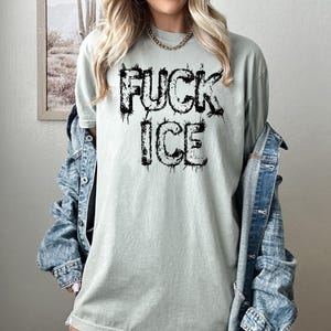 May include: A light green t-shirt with the words "FUCK ICE" printed in black, dripping font. The shirt is a relaxed fit and is being worn with a denim jacket.