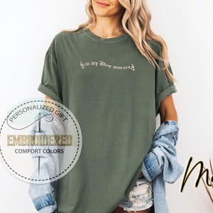 May include: A green t-shirt with the text "Live in my Disney mom era" embroidered in white on the front. The shirt is made of a soft, comfortable fabric and has a relaxed fit.