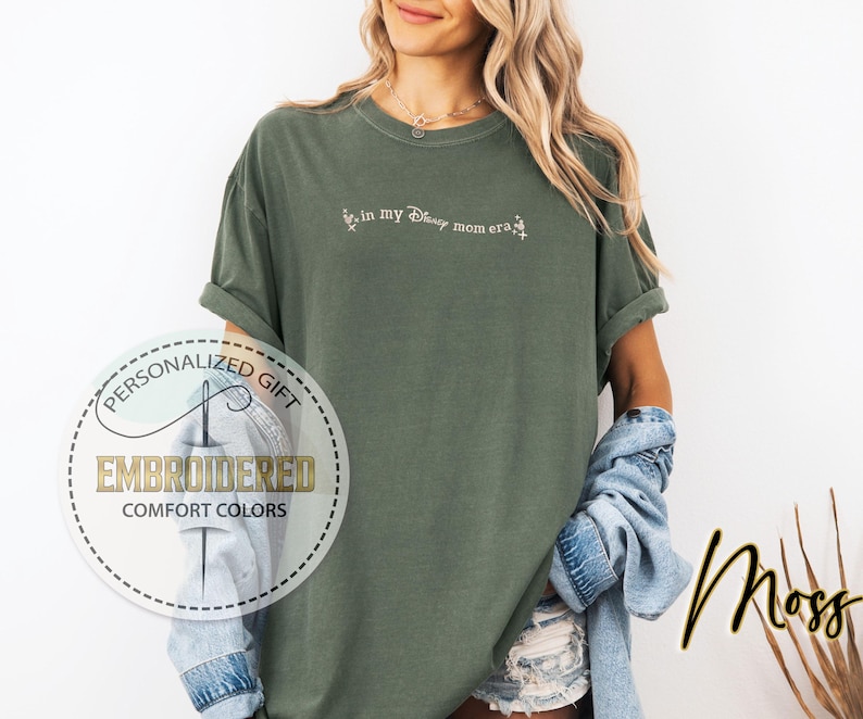 May include: A green t-shirt with the text "Live in my Disney mom era" embroidered in white on the front. The shirt is made of a soft, comfortable fabric and has a relaxed fit.