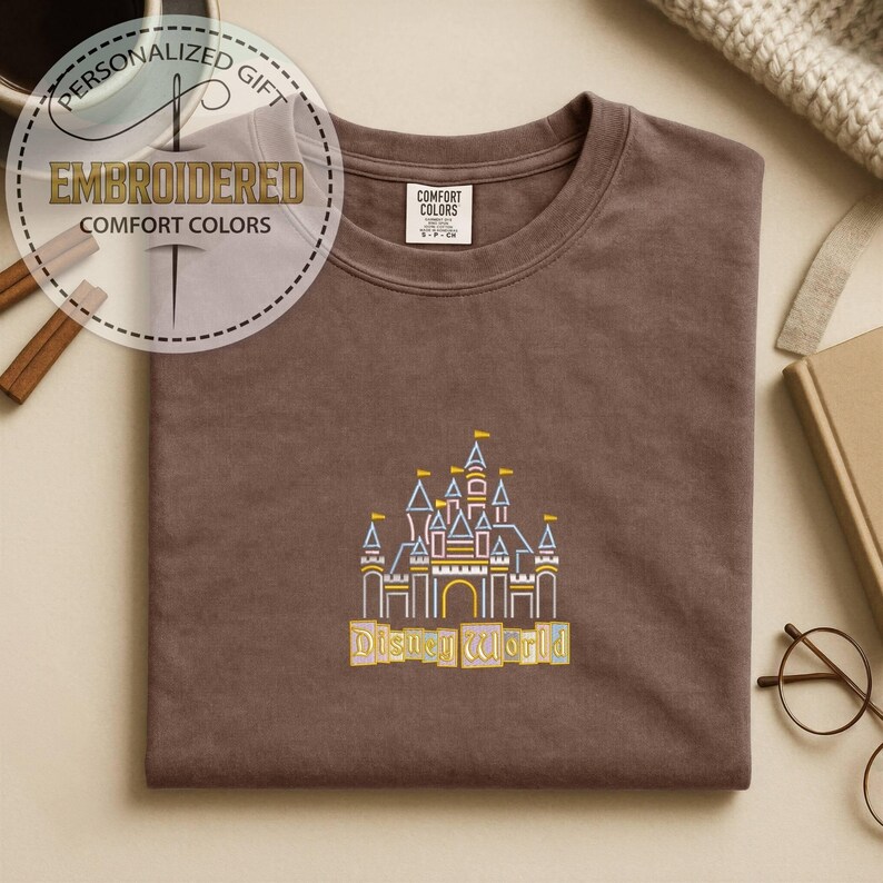 May include: A brown Comfort Colors t-shirt with an embroidered design of a castle and the words "Disney World" in yellow. The shirt has a crew neck and short sleeves. The image also includes text that says "Personalized Gift" and "Embroidered Comfort Colors".