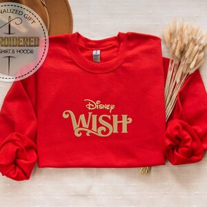 Peut inclure: Un sweat-shirt rouge avec le mot "WISH" en lettres dor&eacute;es, surmont&eacute; du logo Disney. Le sweat-shirt a des manches longues et un col rond. L'image comprend &eacute;galement un chapeau et un bouquet de fleurs s&eacute;ch&eacute;es.