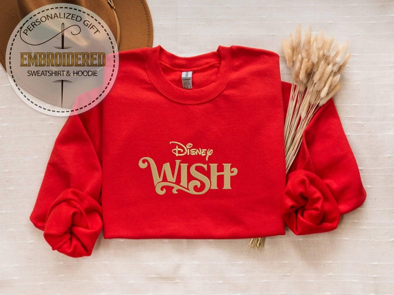 Peut inclure: Un sweat-shirt rouge avec le mot "WISH" en lettres dor&eacute;es, surmont&eacute; du logo Disney. Le sweat-shirt a des manches longues et un col rond. L'image comprend &eacute;galement un chapeau et un bouquet de fleurs s&eacute;ch&eacute;es.