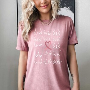 May include: A pink t-shirt featuring a white graphic of different breast shapes and sizes, including a heart with a pink ribbon. The design represents breast cancer awareness.