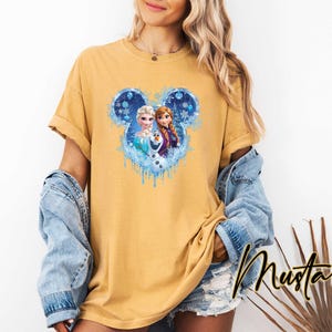 Puede incluir: Camiseta color mostaza con un gr&aacute;fico de Elsa, Anna y Olaf de Frozen, dentro de un dise&ntilde;o en forma de Mickey Mouse con detalles azules. La camiseta tiene mangas cortas y un corte holgado. La palabra "Mustard" est&aacute; escrita en cursiva en la parte inferior derecha.