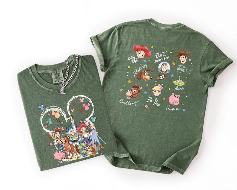 May include: Two olive green t-shirts with Disney's Toy Story characters. One shirt features a Mickey Mouse head design with characters, the other has character portraits and names. A silver chain necklace rests on the first shirt.