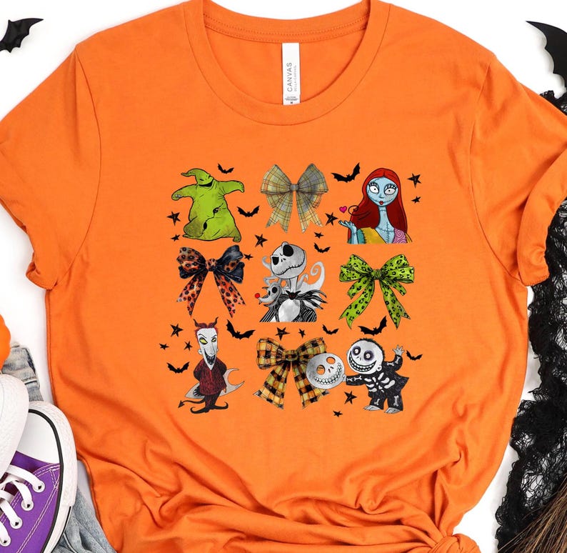 May include: Orange t-shirt featuring a grid of cartoon characters and bows. The characters include a green monster, a skeleton, and a rag doll. The bows have various patterns, and the shirt is decorated with bats and stars.