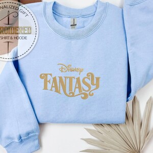 Peut inclure: Sweat-shirt bleu clair avec l'inscription dor&eacute;e "Disney Fantasy". Le sweat-shirt a un col rond et des manches longues. L'image comprend &eacute;galement un logo qui dit "Cadeau Personnalis&eacute; Sweat-shirt & Sweat &agrave; capuche brod&eacute;".