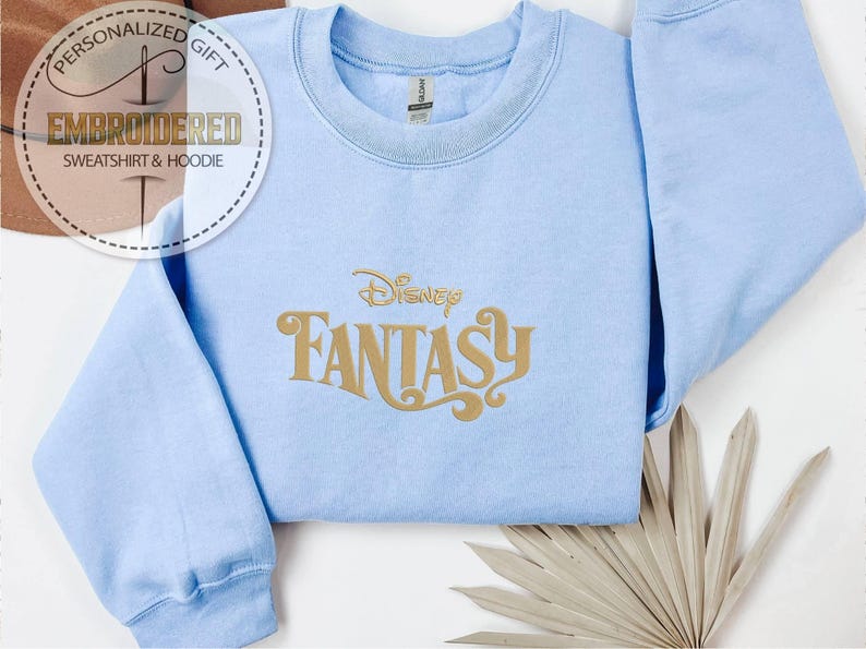Peut inclure: Sweat-shirt bleu clair avec l'inscription dor&eacute;e "Disney Fantasy". Le sweat-shirt a un col rond et des manches longues. L'image comprend &eacute;galement un logo qui dit "Cadeau Personnalis&eacute; Sweat-shirt & Sweat &agrave; capuche brod&eacute;".