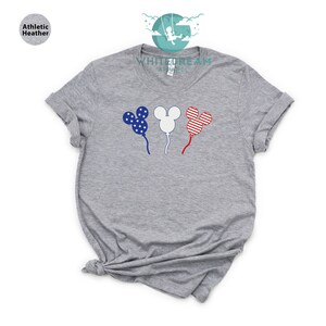 Puede incluir: Una camiseta gris con un dise&ntilde;o de tres globos con forma de cabeza de Mickey Mouse. Los globos est&aacute;n decorados con los colores de la bandera estadounidense: rojo, blanco y azul.