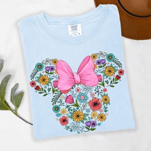 Op de afbeelding: Lichtblauw T-shirt met een bloemen Minnie Mouse-ontwerp. Het ontwerp bevat een roze strik en een verscheidenheid aan kleurrijke bloemen. Het shirt is gemaakt van een zacht materiaal, ideaal voor een casual look.