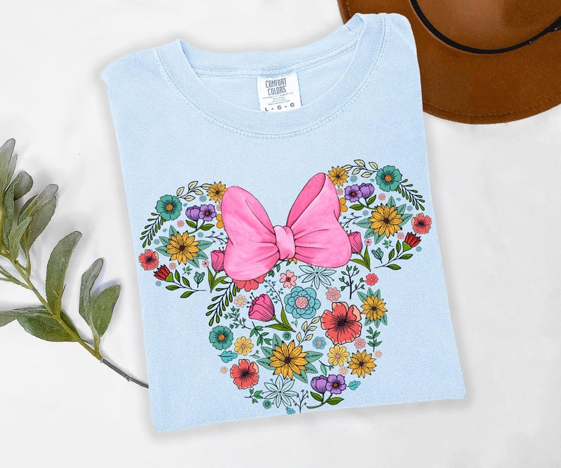 Op de afbeelding: Lichtblauw T-shirt met een bloemen Minnie Mouse-ontwerp. Het ontwerp bevat een roze strik en een verscheidenheid aan kleurrijke bloemen. Het shirt is gemaakt van een zacht materiaal, ideaal voor een casual look.