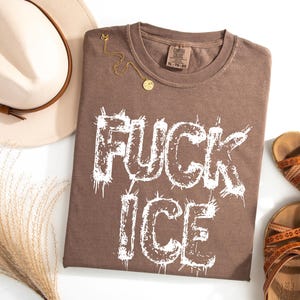 May include: A brown t-shirt with the words "FUCK ICE" in white, a gold necklace, a beige hat with a brown band, and brown sandals. The t-shirt has a crew neck and a tag that reads XL-TG-EG.