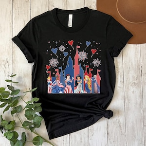 K&ouml;nnte beinhalten: Schwarzes T-Shirt mit einem patriotischen Design mit einer roten und blauen Schloss-Silhouette, Feuerwerk und herzf&ouml;rmigen Ballons. Das Shirt zeigt mehrere Prinzessinnen in rot, wei&szlig; und blauen Kleidern.