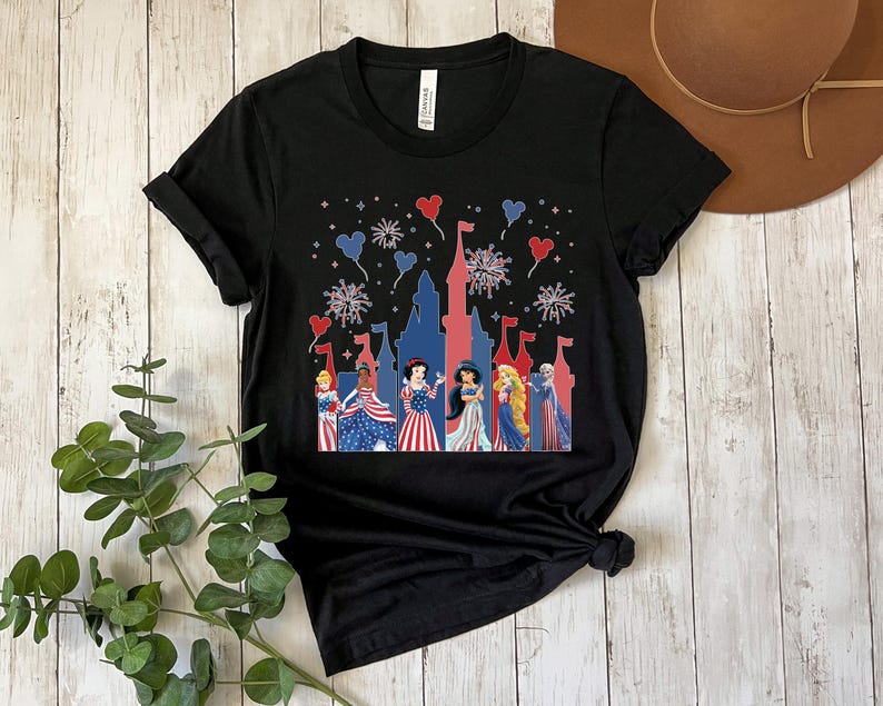K&ouml;nnte beinhalten: Schwarzes T-Shirt mit einem patriotischen Design mit einer roten und blauen Schloss-Silhouette, Feuerwerk und herzf&ouml;rmigen Ballons. Das Shirt zeigt mehrere Prinzessinnen in rot, wei&szlig; und blauen Kleidern.
