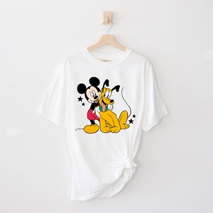 May include: White t-shirt with a cartoon image of Mickey Mouse and Pluto. Mickey Mouse is wearing red shorts and Pluto is wearing a green collar.