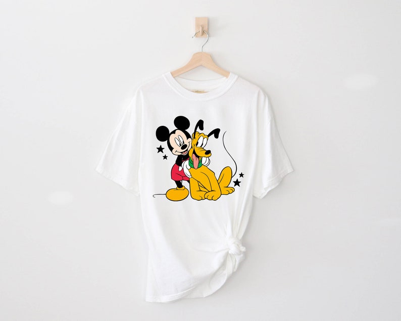 May include: White t-shirt with a cartoon image of Mickey Mouse and Pluto. Mickey Mouse is wearing red shorts and Pluto is wearing a green collar.