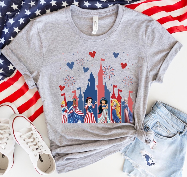 K&ouml;nnte beinhalten: Ein graues T-Shirt mit einem patriotischen Design, das eine Schlosssilhouette, Feuerwerk und Disney-Prinzessinnen in rot, wei&szlig; und blauen Kleidern zeigt. Das Shirt wird mit Jeansshorts und wei&szlig;en Turnschuhen kombiniert, mit einer amerikanischen Flagge im Hintergrund.