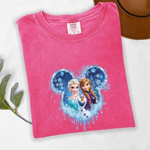 Puede incluir: Una camiseta rosa Comfort Colors con cuello redondo, que presenta un dise&ntilde;o de Elsa, Anna y Olaf de la pel&iacute;cula Frozen, dentro de un dise&ntilde;o en forma de Mickey Mouse. La etiqueta dice L-G-G.