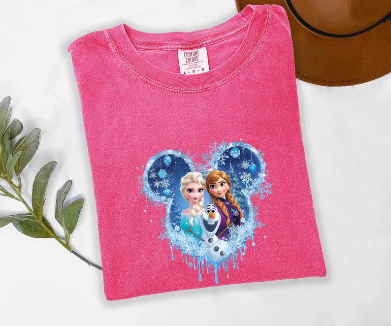 Puede incluir: Una camiseta rosa Comfort Colors con cuello redondo, que presenta un dise&ntilde;o de Elsa, Anna y Olaf de la pel&iacute;cula Frozen, dentro de un dise&ntilde;o en forma de Mickey Mouse. La etiqueta dice L-G-G.