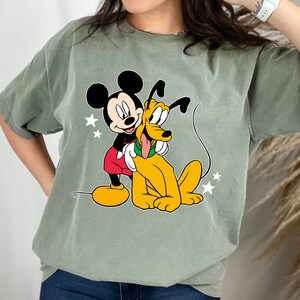 May include: A green t-shirt with a cartoon image of Mickey Mouse hugging Pluto. Mickey is wearing red shorts and yellow shoes. Pluto is yellow with a red collar.