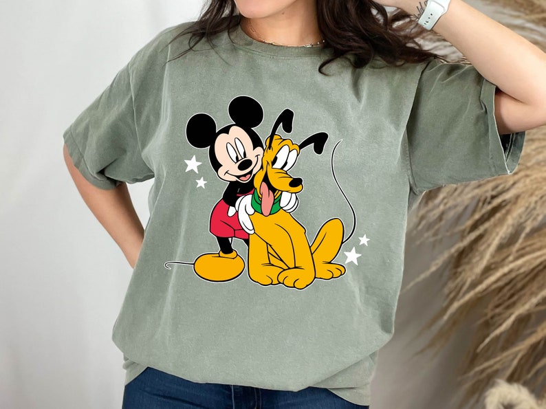 May include: A green t-shirt with a cartoon image of Mickey Mouse hugging Pluto. Mickey is wearing red shorts and yellow shoes. Pluto is yellow with a red collar.