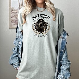 Puede incluir: Camiseta verde claro con el texto "ONYX STORM" sobre un gr&aacute;fico de un drag&oacute;n dentro de un diamante, rodeado por una corona de laurel. El texto "BRAVE THE DARK" est&aacute; debajo. La camiseta es de un material suave.