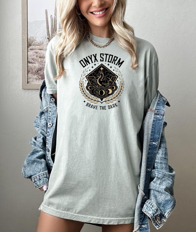 Puede incluir: Camiseta verde claro con el texto "ONYX STORM" sobre un gr&aacute;fico de un drag&oacute;n dentro de un diamante, rodeado por una corona de laurel. El texto "BRAVE THE DARK" est&aacute; debajo. La camiseta es de un material suave.