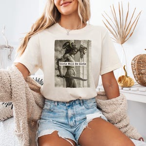 May include: Cream-colored t-shirt featuring a black and white image of a statue holding a sword, with the text "Girls Will Be Girls". The shirt is paired with denim shorts. A cozy, cream-colored cardigan is draped over the arms.