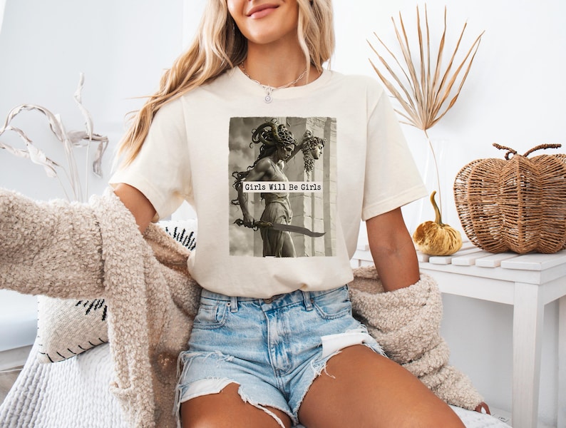May include: Cream-colored t-shirt featuring a black and white image of a statue holding a sword, with the text "Girls Will Be Girls". The shirt is paired with denim shorts. A cozy, cream-colored cardigan is draped over the arms.